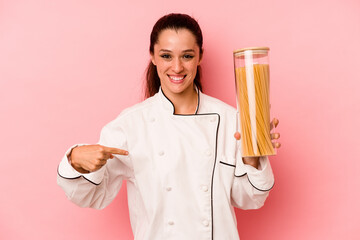 Wall Mural - Young chef caucasian woman holding a spaghettis jar isolated on pink background person pointing by hand to a shirt copy space, proud and confident