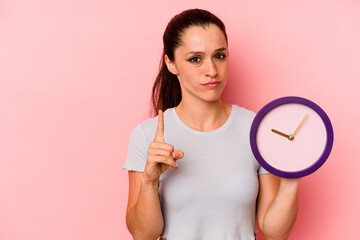 Wall Mural - Young caucasian woman holding a clock isolated on pink background showing number one with finger.