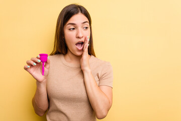 Young caucasian woman holding a menstrual cup isolated on yellow background is saying a secret hot braking news and looking aside