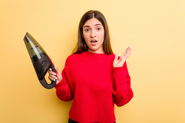 Wall Mural - Young caucasian woman holding a hand vacuum cleaner isolated on yellow background surprised and shocked.