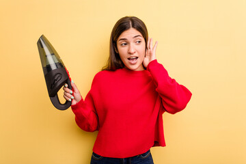 Wall Mural - Young caucasian woman holding a hand vacuum cleaner isolated on yellow background trying to listening a gossip.