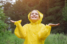 Joyful Fair-haired Teenager Girl Stands In Rain On Forest Glade. Smiling Girl In Yellow Raincoat With Hood Enjoys Catching Raindrops With Hands
