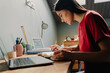 © Drobot Dean - Young woman writing down notes while working on laptop at home