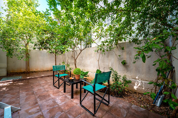 Backyard terrace with tiled floor and garden furniture, two green chairs and smal black table with decor on it, trees and white wall around. Summer sunny day. 