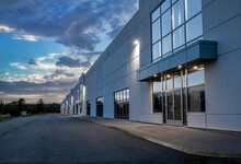 Exterior View Of A Generic Business Park Building At Dusk With Multiple High Bay Units, Prefab Metal Cladding, Glazing, Aluminum Mullions, Exterior Lights On, Nobody