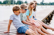 © Iryna - Four cute different age boys and girls sitting on a wooden platform by the river or lake dipping their feet in the water on warm summer day. Outdoor activities for kids concept.