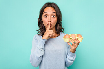 Wall Mural - Young hispanic woman holding eggs isolated on blue background keeping a secret or asking for silence.