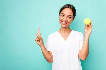 Wall Mural - Young hispanic physiotherapy holding a tennis ball isolated on blue background showing number one with finger.