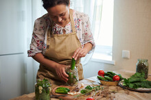 Charming Woman, Housewife Puts Freshly Picked Organic Cucumbers In A Glass Jar For Preservation, Before Pouring The Brine While Preparing Homemade Pickles, Stands At The Table With Various Ingredients