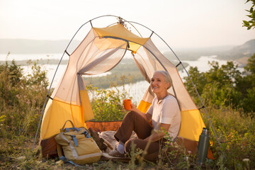 portrait of a smiling senior woman relaxed in a tent and drinks tea outdoors at sunset. copy space. Slow life. Enjoying the little things. spends time in nature in summer