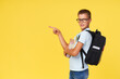 Portrait of a schoolboy in glasses with textbooks and a backpack on a yellow background pointing to the left. Back to school