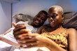 © Ezequiel Giménez/Stocksy - Black woman using smartphone near boyfriend on bed