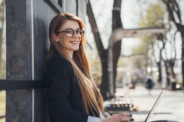 Red haired woman sitting in the park and posing while working on her online project. Online job concept. Woman working outdoors in nature. Mobile Office