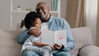 © Yuliia - Mature african american man with cute kid girl hugging sitting in room on sofa father reads greeting card loving daughter made gift with own hands for birthday or father's day dad happy cuddling baby