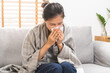 © KMPZZZ - Sick, Coronavirus covid-19 asian young woman, girl headache under blanket have a fever, flu and use tissues paper sneezing nose, runny sitting on sofa bed at home. Health care on virus person.