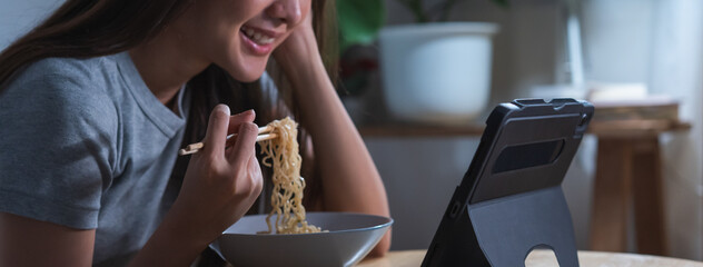 Relaxation, smile asian young woman hand use chopstick eating instant ramen, noodles while using, watching video, movie, media on pc, tablet and browsing internet in overtime night, late time at home.