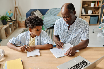 Wall Mural - African tutor pointing at copybook and explaining ne material to pupil at table at home schooling