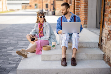 Stylish colleagues work on laptop and eat take away food while sitting and talking together on a street at office district. Concept of modern business lifestyle