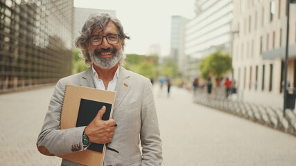 Smiling mature businessman with beard in eyeglasses wearing gray jacket looks around and walks down the street past modern buildings