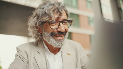 Mature businessman with beard in eyeglasses wearing gray jacket sits in cafe. Middle aged manager having video conference, video call on laptop pc computer sit at cafe outdoor