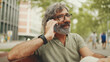 © Andrii Nekrasov - Laughing middle-aged man with gray hair and beard wearing casual clothes sits on bench and uses mobile phone. Mature gentleman in eyeglasses talking on smartphone