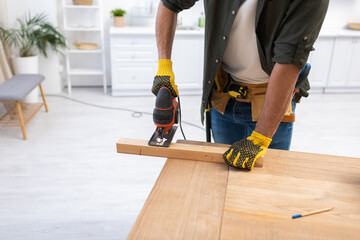 Wall Mural - Cropped view of man in gloves holding jigsaw machine while sawing plank at home