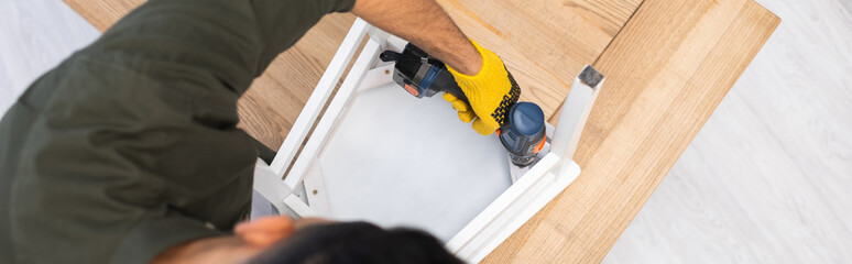 Wall Mural - Overhead view of young man repairing chair with electric screwdriver at home, banner