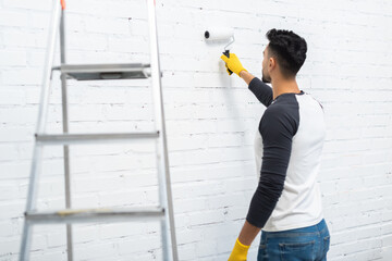 Wall Mural - Arabian man in gloves coloring wall near ladder at home