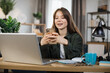 © sofiko14 - Confident young woman writing message during mobile conversation at office. Caucasian female worker with long dark hair sitting at table with opened laptop.
