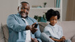 © Yuliia - Close-up happy african american father and daughter laughing watching funny tv show movie relaxing on couch man holding remote control showing thumb up gesture of approval dad and kid girl having fun