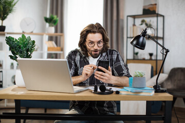 Wall Mural - Young bearded excited man in casual outfit typing on mobile phone while sitting at workplace with laptop. Concept of corporate communication and good news.