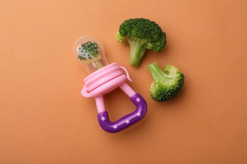 Wall Mural - Nibbler with boiled broccoli on pale orange background, flat lay. Baby feeder