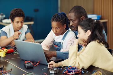 Wall Mural - Portrait of black teenage girl using laptop in school during engineering class with male teacher and diverse group of children
