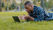 © Yuliia - African american guy young business man user lying on green grass lawn in park using laptop online call conference chat remote conversation distant talk talking speaking with friends family outdoors