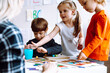 © Татьяна Волкова - Two beautiful little girls standing at table with pencils in bright classroom. Little boy sitting, playing board games.