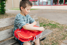 The Boy Sits On A Bench In The School Yard And Takes Out A Lunch Box From A School Backpack. Children Have Snacks Between Classes.