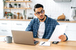 © Kateryna - Focused serious arabian or indian guy with glasses, freelancer or office worker working remotely, sit at a desk at home in kitchen, working on a project uses laptop, concentrated analyzing documents
