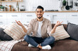 Relaxation and meditation concept. Happy peaceful relaxed indian or arabian man in casual clothes, sitting alone at home in living room on the couch and meditating in the lotus position, smiling