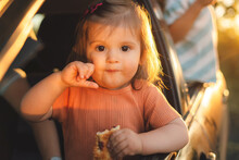Caucasian Baby Girl Enjoying Eating Snack While Stuck Her Head Out The Car Window, Safety Seat During Drive. People Lifestyle Concept. Sweet Food. People