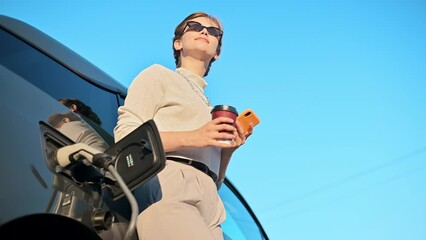 Wall Mural - A young blonde woman with smartphone and coffee at a car charging station with charging electric car nearby in Chisinau, Moldova