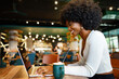 © fotofabrika - Smiling young african woman sitting with laptop in cafe