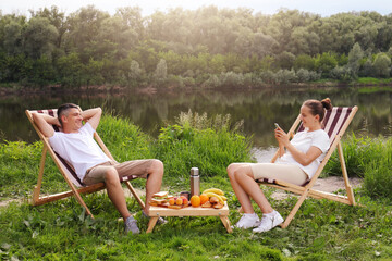 Wall Mural - Outdoor shot of young couple sitting on deck chair and talking near the river, relaxed man sits with hands behind head, woman using cell phone for checking social networks, family relaxing outdoor.