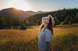 Young beautiful happy cheerful woman in a hat enjoying the sunset in a meadow with mountains view