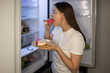 Woman eats a sweet delicious calorie sandwich with cream in her pajamas at night near the refrigerator. Concept: night meal, calories, food during stress and depression, bulimia, eating disorder