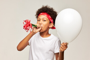 Portrait of funny happy boy kid of african ethnicity blowing birthday horn holding mockup balloon on gray studio background wearing white copy space t-shirt and red bandana. Happy holidays