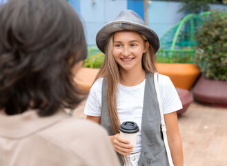 Wall Mural - Smiling girl meeting with female friend holding coffee outdoors. Happy woman wearing hat and waistcoat drinking coffee and talking to girlfriend. Female friendship concept