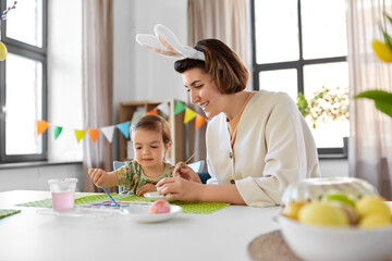 Poster - holidays, family and people concept - happy mother and little baby daughter coloring easter eggs with watercolors at home