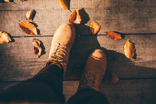 Tourist In Beige Boots Walks Along Timber Deck Bridge Covered With Yellow Leaves In Picturesque Autumn Park.