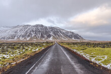 A Moss-covered Landscape Of Jumbled Lava Borders This Narrow Road In Iceland. The Road Continues On Toward A Snow Dusted Mountain Ahead.