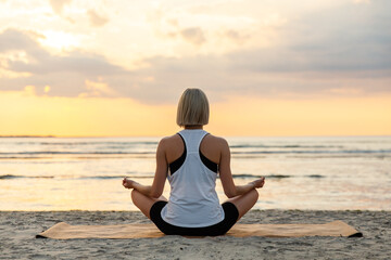 Wall Mural - yoga, mindfulness and meditation concept - woman meditating in lotus pose on beach over sunset